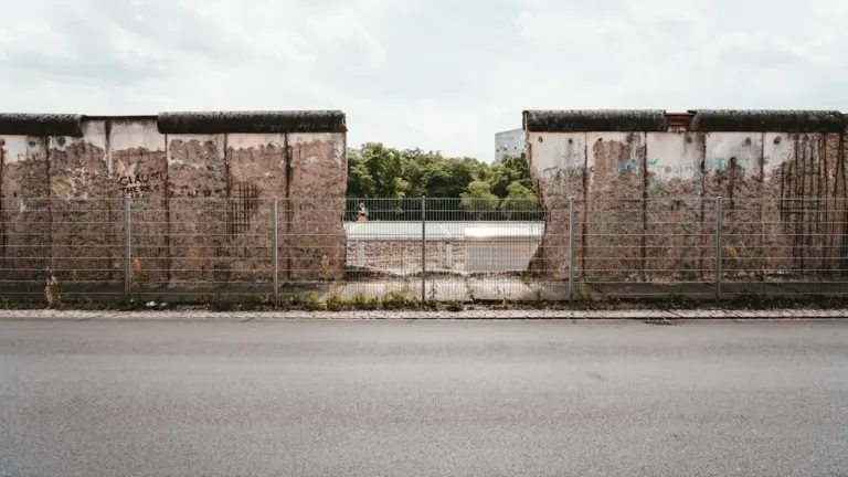 Ein grauer Metallzaun vor den Überresten der Berliner Mauer bei Tageslicht.