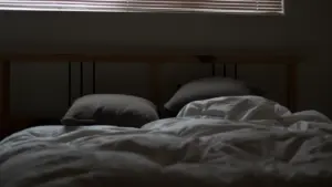 Ein ungemachtes Bett mit geschlossenen Jalousien bei abgedunkelter Atmosphäre im Schlafzimmer.