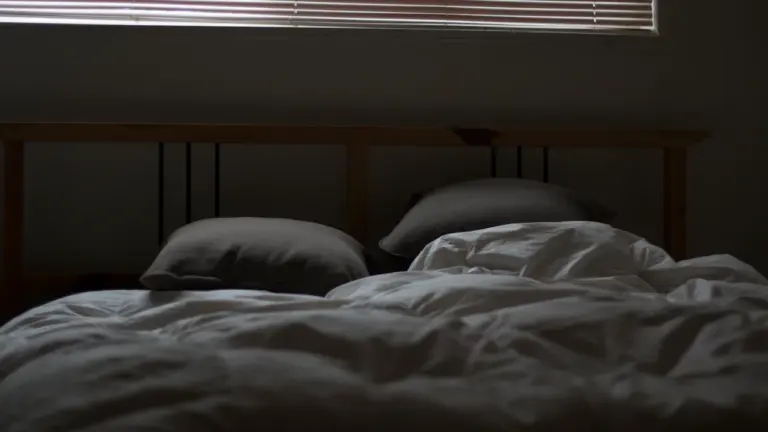 Ein ungemachtes Bett mit geschlossenen Jalousien bei abgedunkelter Atmosphäre im Schlafzimmer.