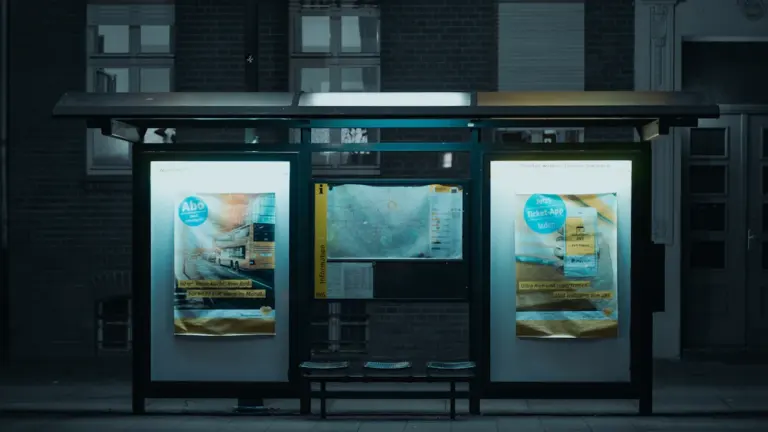 Eine nächtliche Bushaltestelle in einer Stadt mit beleuchteten Plakaten und Sitzgelegenheiten.