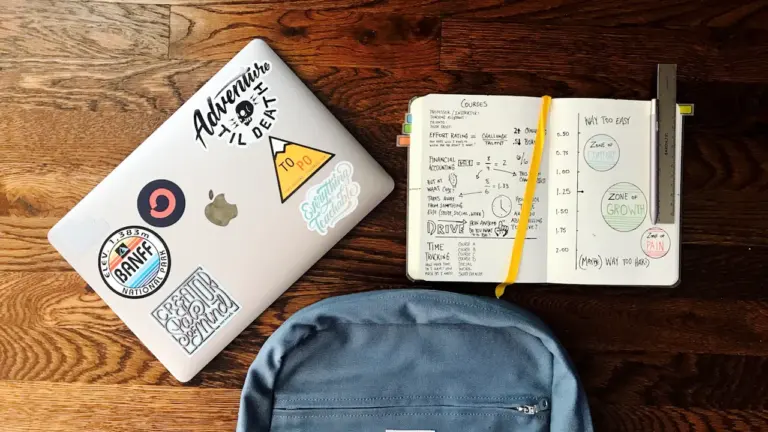 Blauer Rucksack neben einem offenen Notizbuch und Laptop auf Holzboden für den Schulstart.