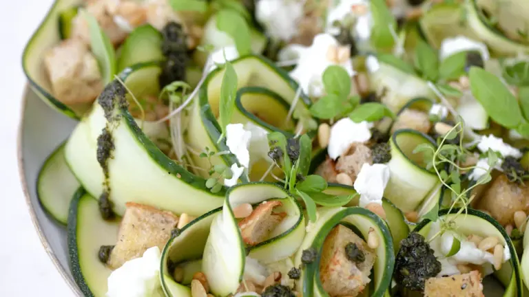 Nahaufnahme eines sommerlichen Zucchini-Salats mit Feta, Kräutern und Pinienkernen auf einem Teller.
