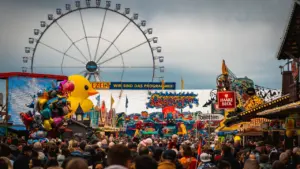 Ein belebter Jahrmarkt vor einem großen Riesenrad unter einem bewölkten, regnerischen Himmel.