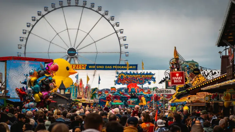 Ein belebter Jahrmarkt vor einem großen Riesenrad unter einem bewölkten, regnerischen Himmel.